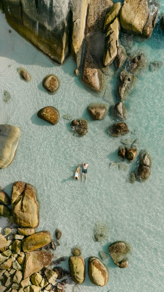 Aerial view of a person and a dog lying on clear, shallow water surrounded by large rocks and boulders at a beach.