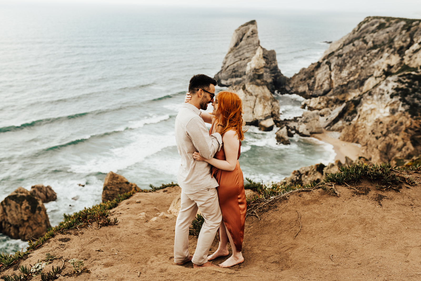 man and woman embracing eachother on the cliffs of Praia da Ursa woman is wearing coral silk dress and has red hair