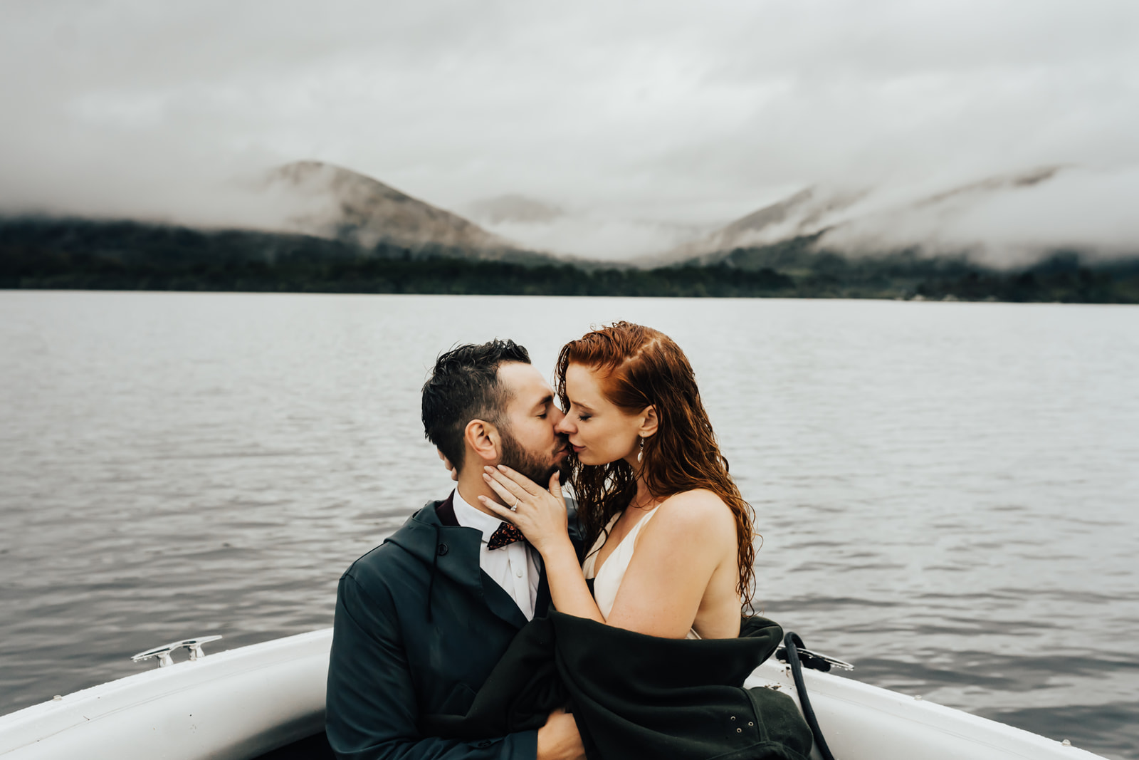 bride and groom kissing on a boat infront of mountains covered in mist