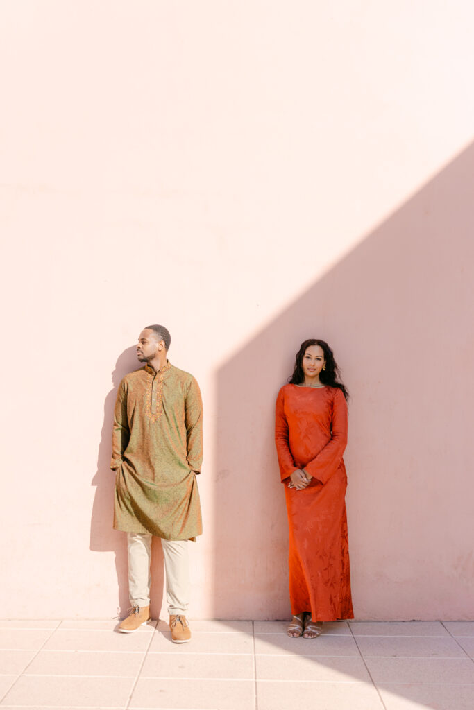 A man and woman stand against a pale pink wall, casting shadows. The man wears a green kurta and beige pants, looking left. The woman wears a long, bright red dress and looks forward. Sunlight creates a diagonal shadow on the wall.