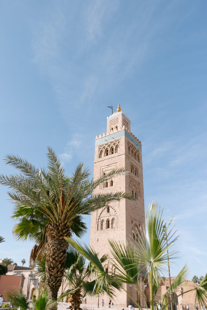 A tall, historic stone minaret stands against a blue sky, surrounded by palm trees. The building features intricate designs and a gold finial at the top.