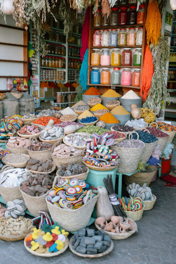 Colorful market stall displays baskets of spices, herbs, and vibrant textiles. Glass jars of pigments and spices fill shelves in the background, while woven baskets hold spices, soaps, and decorative items on tables.