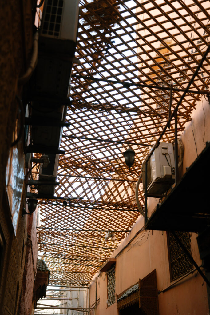 A narrow alleyway with walls painted in warm tones, covered by a woven bamboo lattice roof letting sunlight filter through, with air conditioning units and lamps attached to the walls.