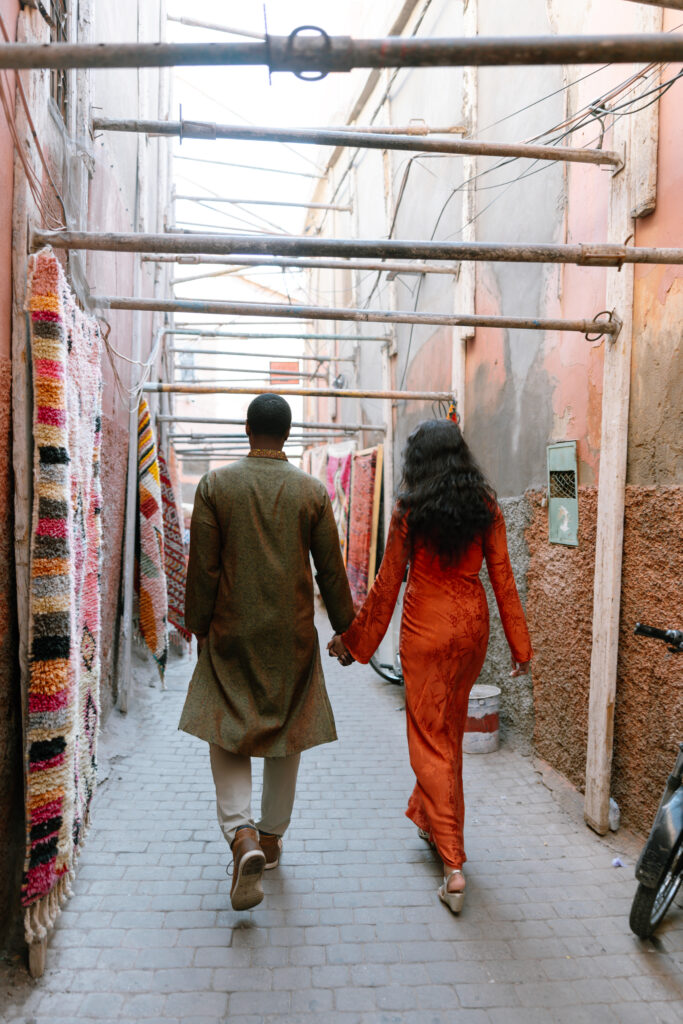 A couple walks hand in hand down a narrow, colorful alley lined with hanging rugs and textiles, with a bicycle parked nearby.