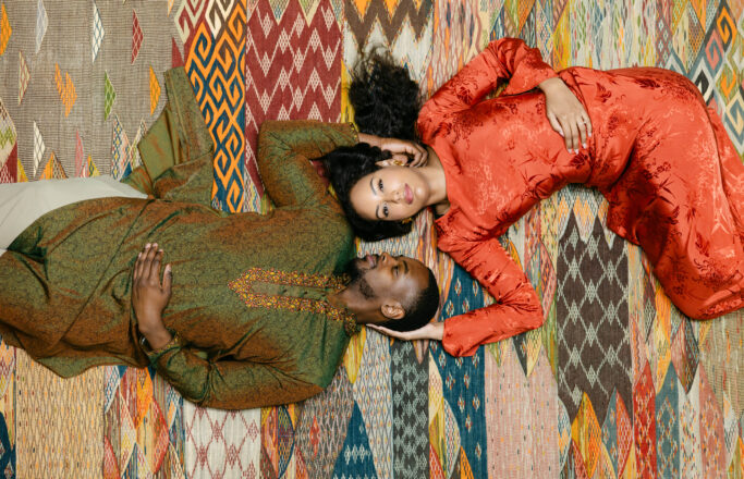 A man and woman in traditional, colorful attire lie on a patterned rug. The man wears green, the woman wears red, and they face upward, heads close together, with the mans hand gently holding her hair.