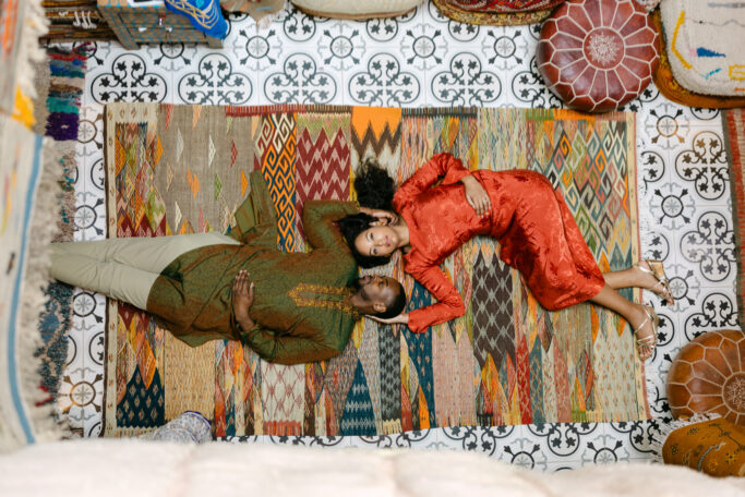 A couple lies on a colorful patterned rug, facing each other. The woman in a red dress rests her head on the man’s arm. The scene is surrounded by patterned cushions and intricate black-and-white floor tiles.