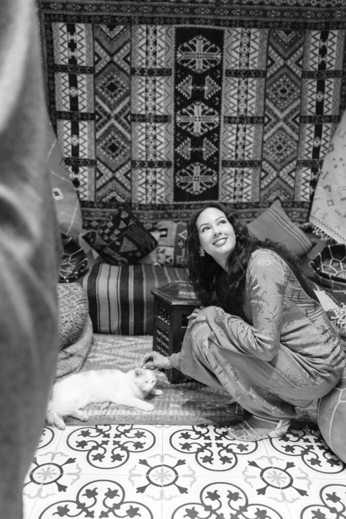 A smiling woman in traditional clothing sits on a patterned floor beside a small white dog, with patterned rugs and textiles hanging in the background. The scene has a cozy, bohemian atmosphere.