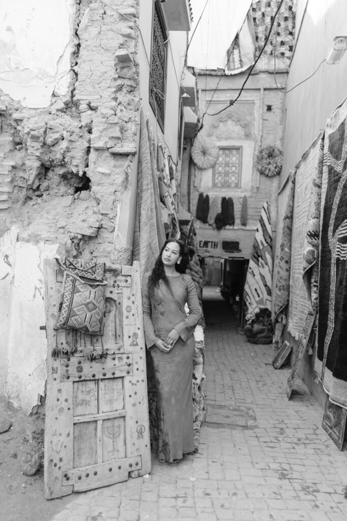 A woman in a long dress leans against a worn, rustic doorway in a narrow, cobblestone alleyway lined with hanging rugs and textiles. The scene is in black and white, evoking a historic or traditional market setting.