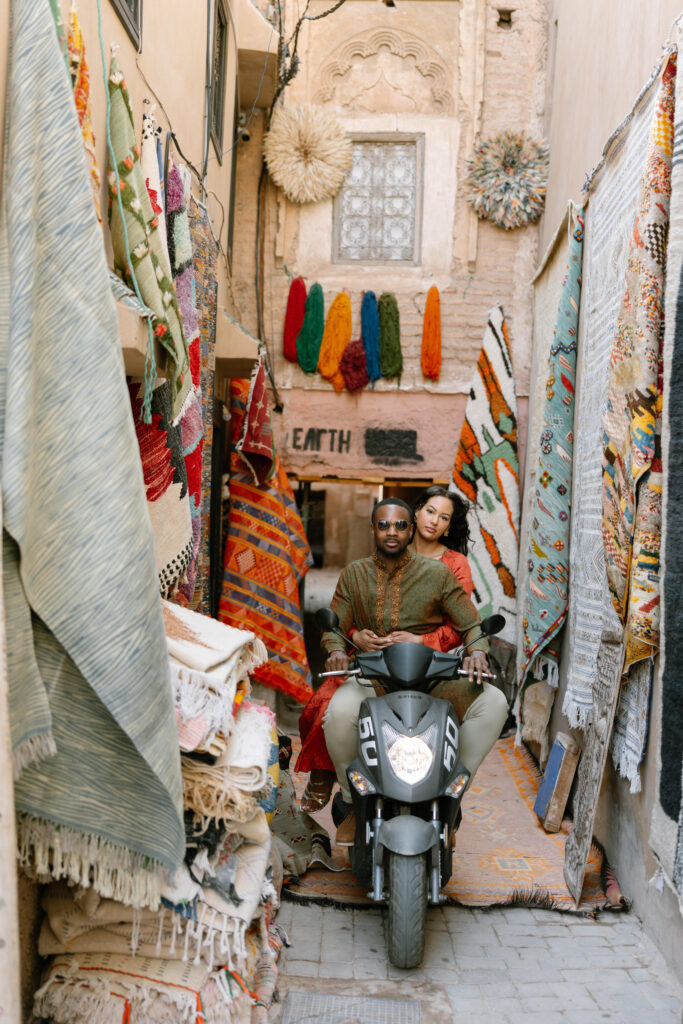A man and woman ride a black scooter through a narrow, colorful alley lined with hanging rugs and textiles. The alley is vibrant and cozy, with patterned fabrics draped on both sides and an ornate window above.
