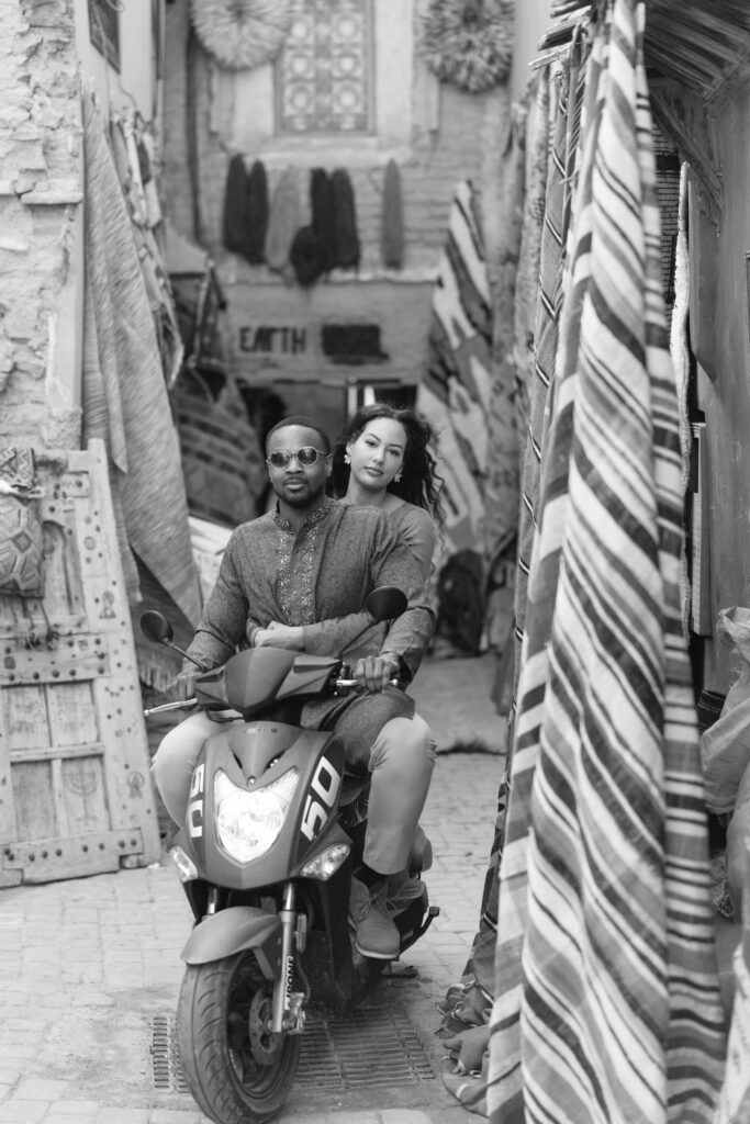 A man and woman ride a scooter down a narrow, vibrant market alley lined with textiles and rugs. The man drives while the woman sits behind him, both wearing casual clothing and sunglasses. The photo is in black and white.