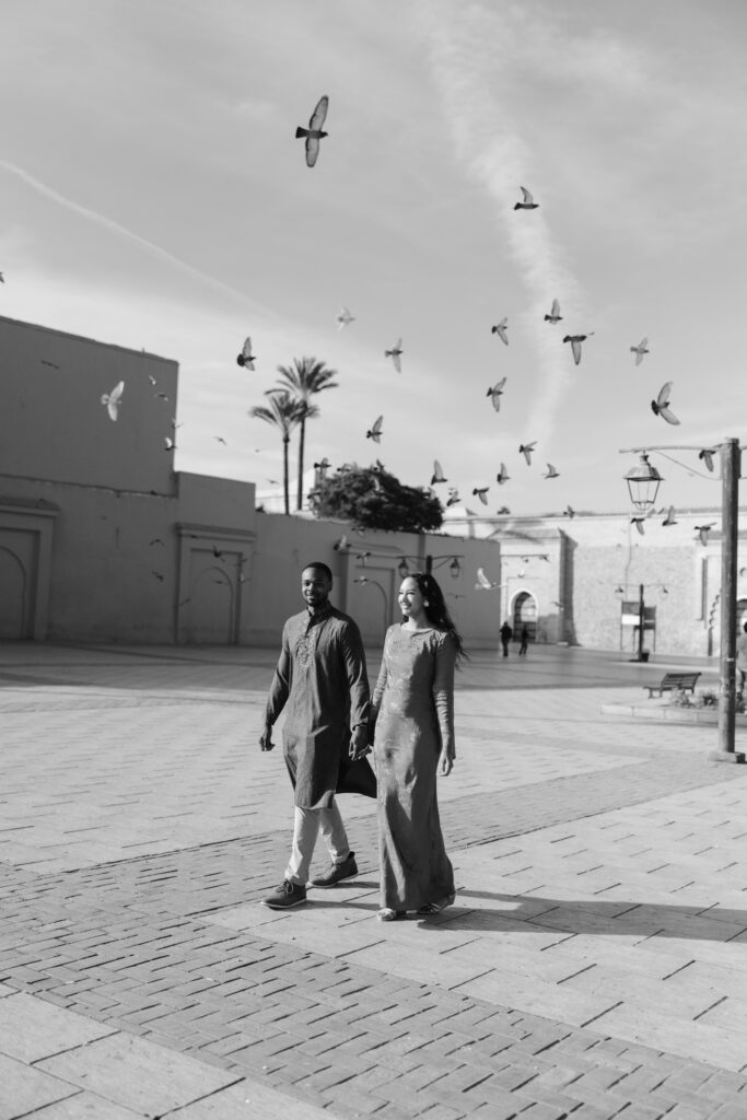 A man and woman in traditional attire walk hand in hand across a spacious, sunlit plaza, with birds flying overhead and tall walls and palm trees in the background. The image is in black and white.