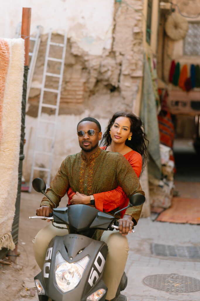 A couple rides a black scooter through a narrow, rustic alleyway. The man drives while the woman sits behind him, holding his waist. Both wear colorful traditional clothing and have a calm expression. Rugs and fabrics hang nearby.