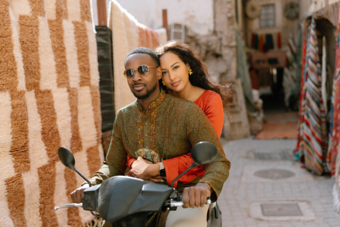 A couple sits together on a scooter in a narrow street lined with colorful rugs. The man wears sunglasses and a patterned shirt, and the woman in an orange top embraces him from behind, both looking at the camera.