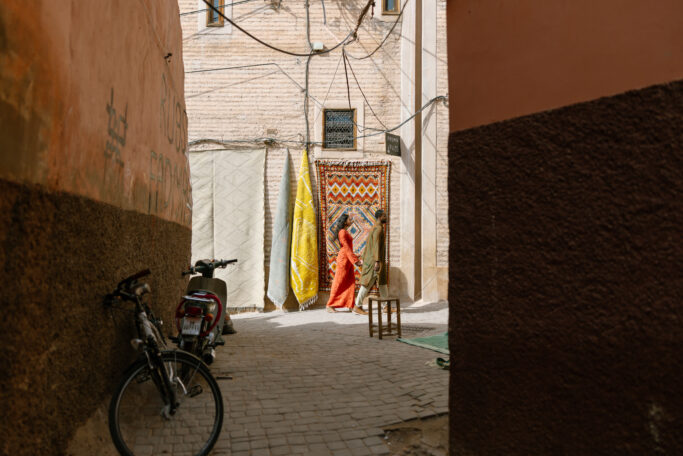 A woman in an orange dress hangs colorful rugs on a wall in a narrow sunlit alleyway, with bicycles and a motorcycle parked nearby. The walls are painted in earthy tones and the ground is cobblestone.