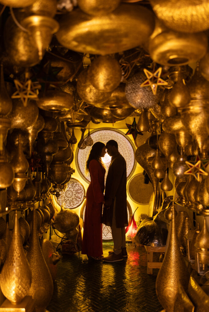 A couple stands face to face, holding hands in a warmly lit room filled with hanging golden lamps and lanterns, creating a romantic and magical atmosphere.