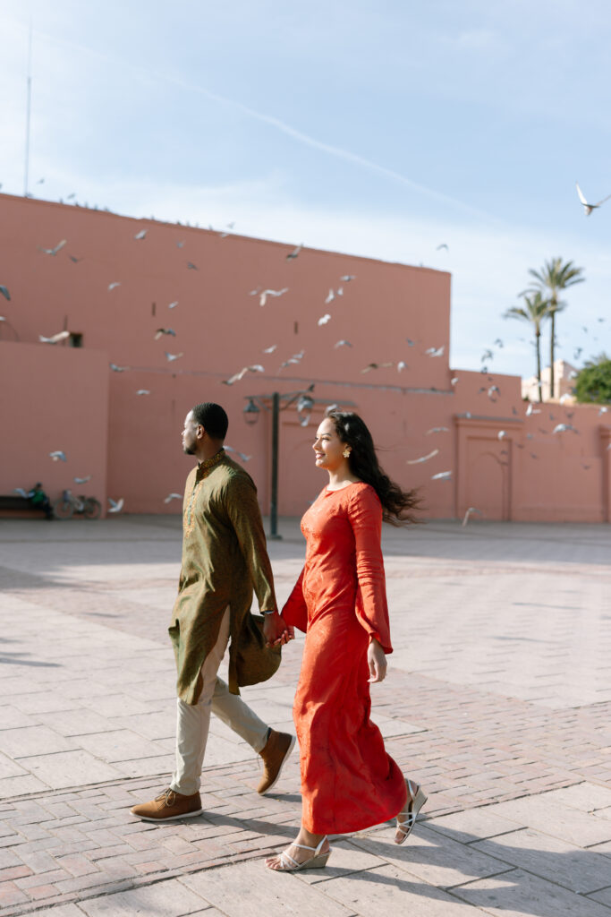 A couple dressed in traditional attire walks hand in hand across a sunlit plaza with peach-colored walls, scattered birds in flight, and palm trees in the background.