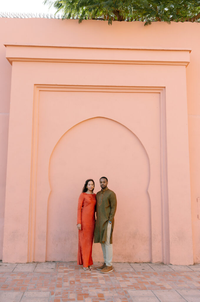 A woman in an orange dress and a man in a green kurta stand side by side in front of a large, pink arched wall with geometric patterns, under a clear sky and leafy branches above.
