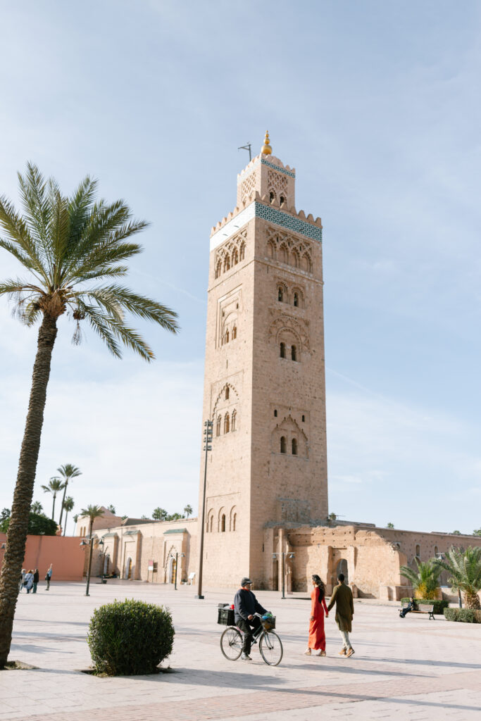 A tall, historic stone tower stands in a sunny square with palm trees. Two people walk together near the tower, while a person rides a bicycle in the foreground. The sky is clear and blue.