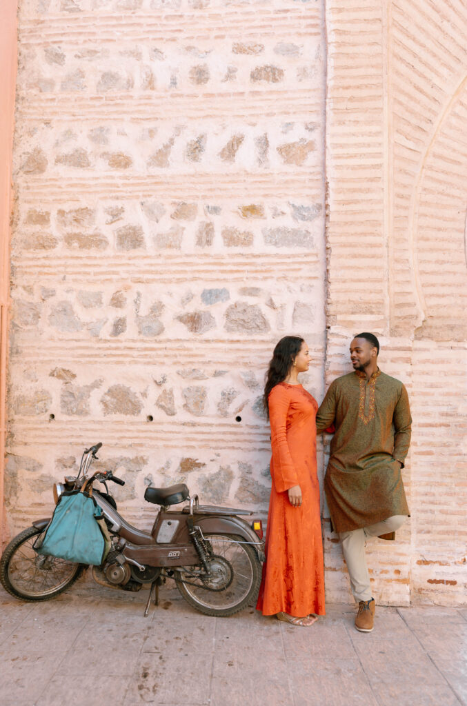 A woman in an orange dress and a man in a green kurta stand close together against a textured stone wall, next to a parked bicycle with a blue bag. They look at each other, appearing relaxed and happy.