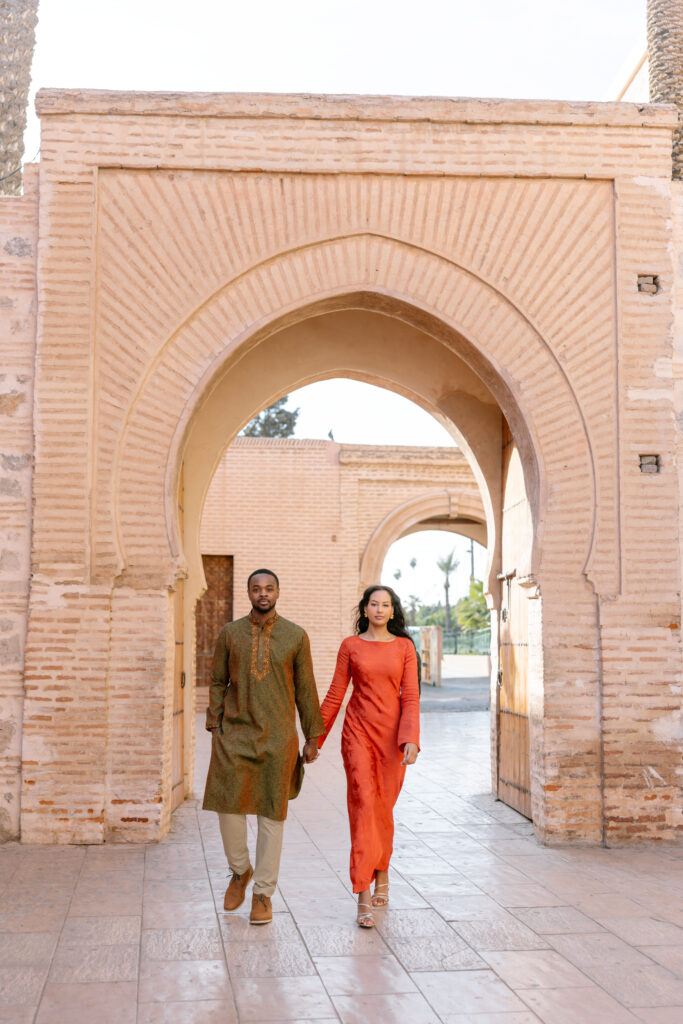 A man and woman holding hands walk through a large, ornate stone archway. The man wears a green traditional outfit, and the woman wears a long red dress. Sunlight falls softly on the scene, highlighting the architectural details.