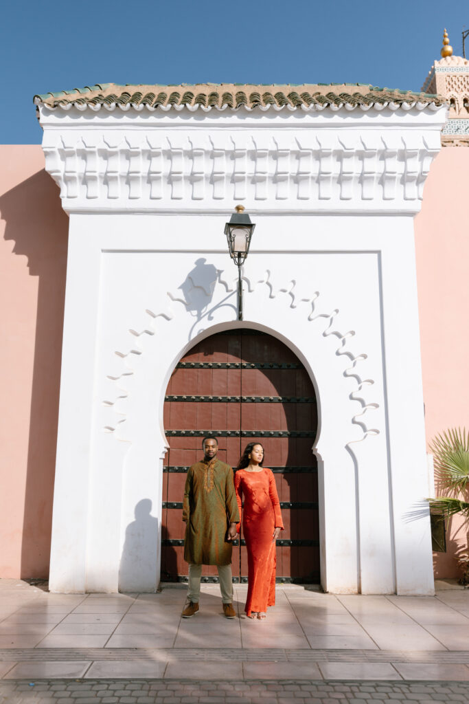 A man and woman stand holding hands in front of a large, ornate white archway with a wooden door and a lantern above, set against a pink wall under a clear blue sky.