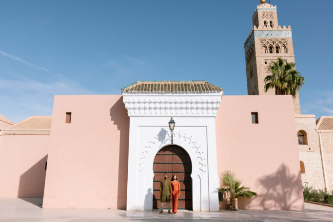 Two people stand in front of a large arched white entrance set in a pink wall, with a tall historic stone tower and palm tree in the background under a clear blue sky.