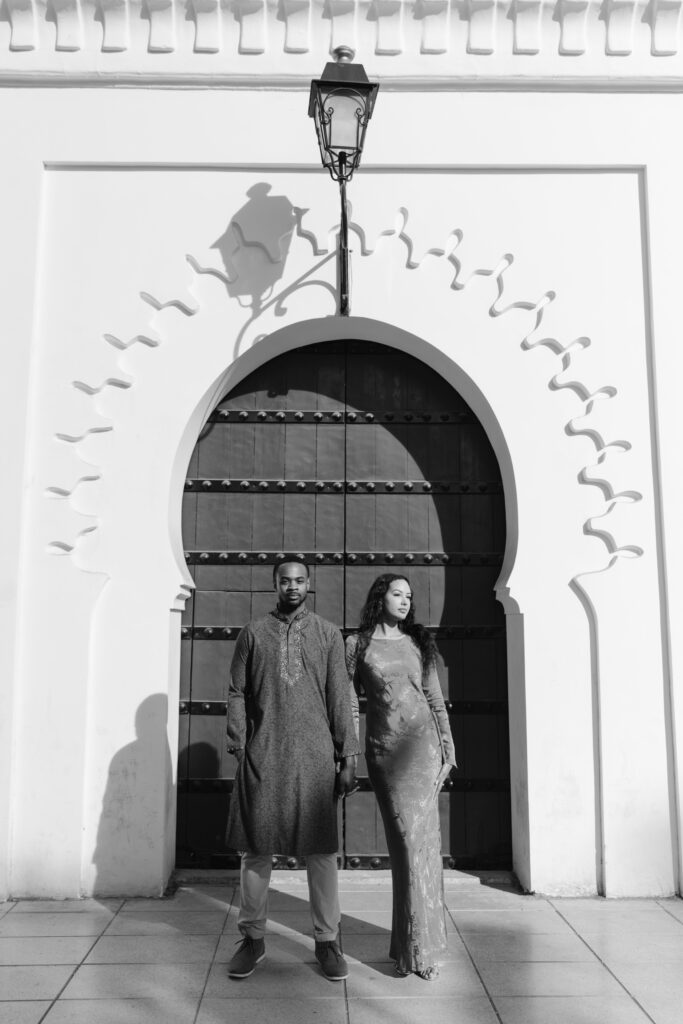 A man and a woman stand side by side in front of an ornate arched doorway with intricate detailing. The shadows and sunlight create contrast on the white wall behind them. Both are dressed in traditional clothing.