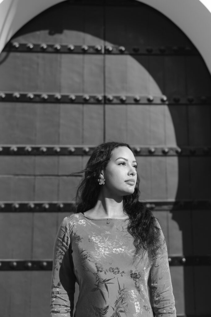 Black and white photo of a woman with long hair standing in front of a large arched door, looking thoughtfully to the side; sunlight creates a shadowed arch on the door behind her.