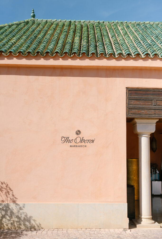 A peach-colored wall with The Oberoi Marrakech and a palm tree logo, featuring a green-tiled roof above and a white column next to a wooden doorway under a clear blue sky.