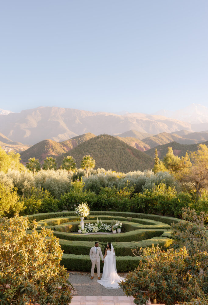 A bride and groom stand hand in hand in the center of an ornate garden maze, surrounded by lush greenery, with mountains and a clear blue sky in the background.
