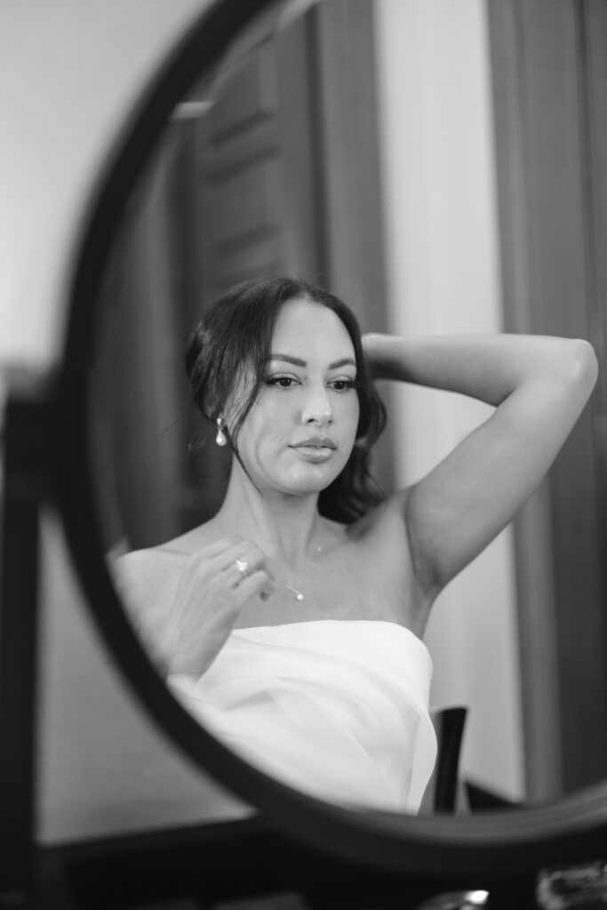 A black-and-white photo shows a woman with dark hair, wearing a strapless top, looking at herself in a round mirror while adjusting her hair and necklace. The background is softly blurred.