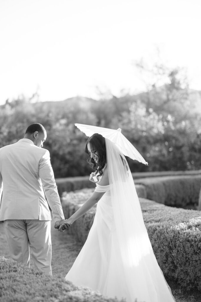 A bride in a flowing gown and veil holds a parasol and smiles back, holding hands with her groom as they walk through a sunlit garden. The photo is in black and white, with soft, romantic lighting.