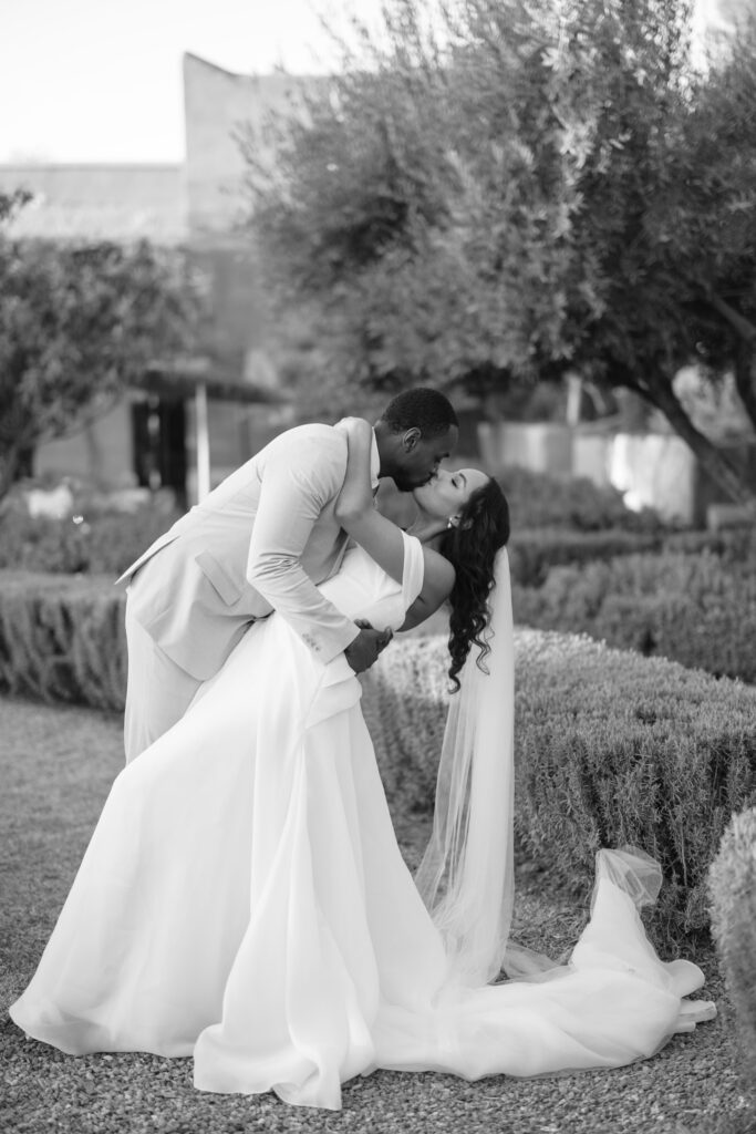 A groom in a light suit dips and kisses his bride, who wears a flowing gown and veil, in a garden setting with manicured bushes and trees. The moment is romantic and joyful. The image is in black and white.