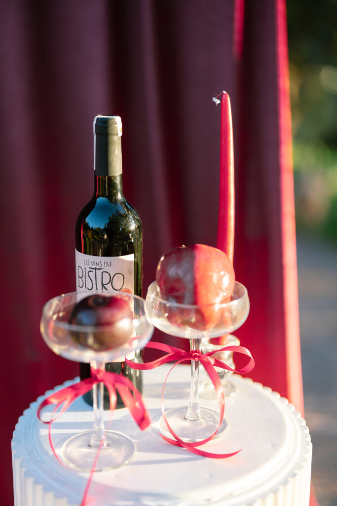 A wine bottle, a red candle, and two apples in glass dessert cups with red ribbons are displayed on a white round table. A deep red curtain is in the background, lit by sunlight.
