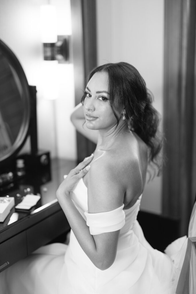 A woman in an off-the-shoulder dress sits at a vanity, looking over her shoulder and smiling softly. Her hair is styled in loose waves, and the scene is elegantly lit, creating a serene and graceful atmosphere.