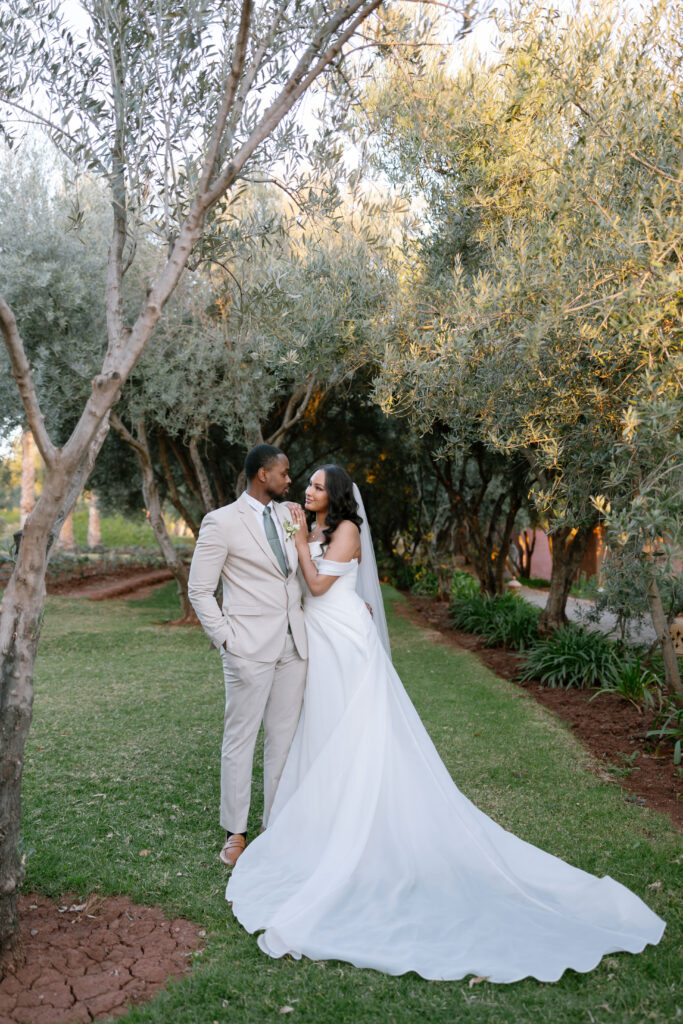 A bride and groom stand outdoors on grass beneath leafy trees, smiling lovingly at each other. The bride wears a long white gown and veil; the groom is in a light beige suit with no tie. Sunlight filters through the trees.