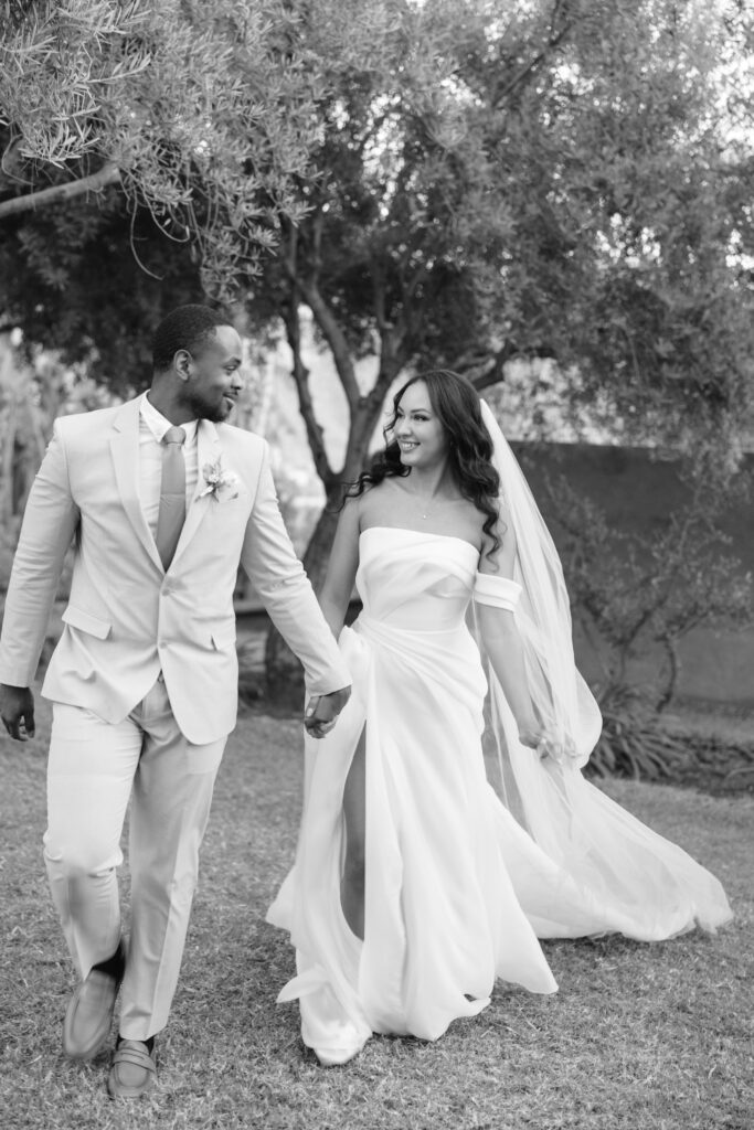 A bride and groom hold hands and smile at each other while walking outdoors. The bride wears an off-the-shoulder gown and veil, and the groom wears a light-colored suit. Trees and grass are visible in the background.
