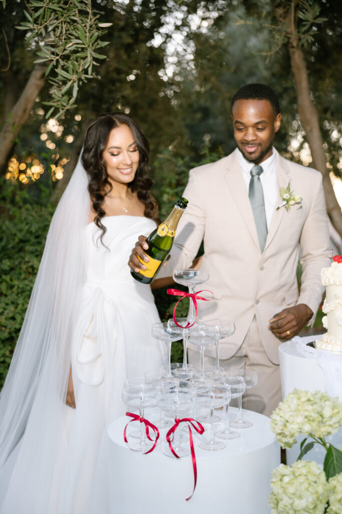 A bride in a white dress and a groom in a beige suit pour champagne into a tower of glasses decorated with red ribbons at an outdoor wedding, surrounded by greenery.