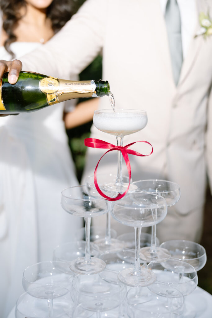 A close-up of champagne being poured into a glass at the top of a pyramid of coupe glasses. The top glass has a red ribbon tied around its stem, and two people in formal attire are partially visible in the background.