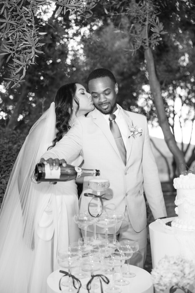A bride in a veil kisses the groom’s cheek as he pours champagne into a tower of glasses at an outdoor wedding, surrounded by trees and a wedding cake.
