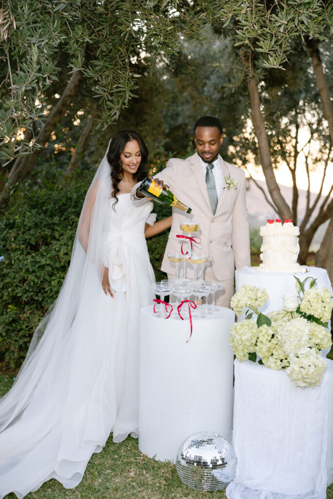 A bride and groom stand outdoors by a champagne tower, with the groom pouring champagne. The bride smiles, wearing a white gown and veil. There are flowers, a wedding cake, and a disco ball nearby.