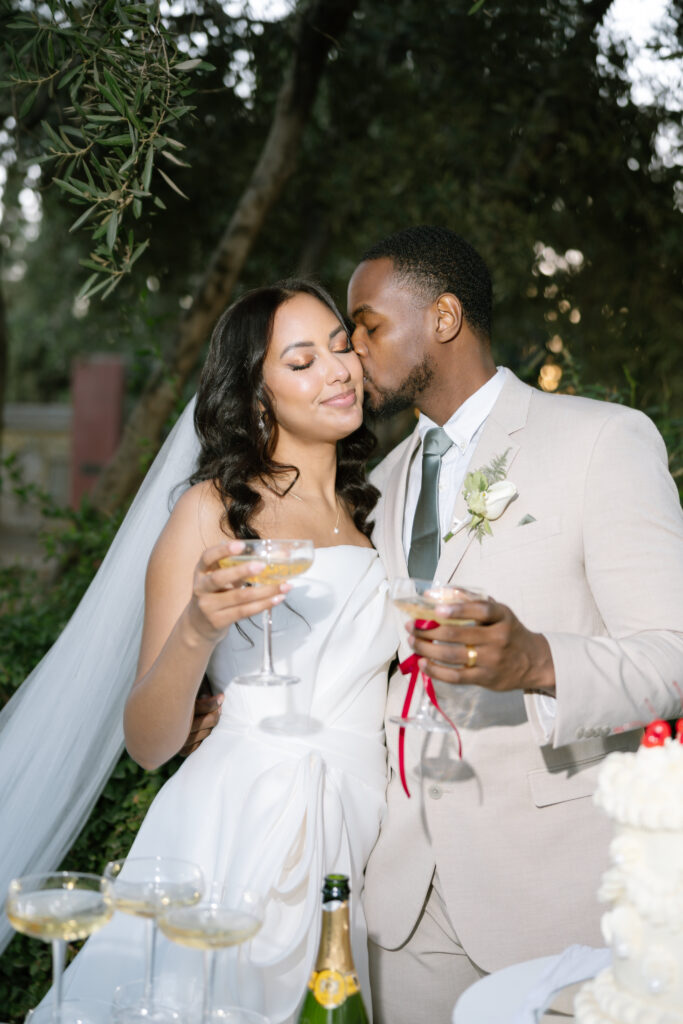A newlywed couple stands close together outdoors, the groom kissing the bride on the forehead as they smile and hold glasses of champagne, dressed in wedding attire, with greenery in the background.