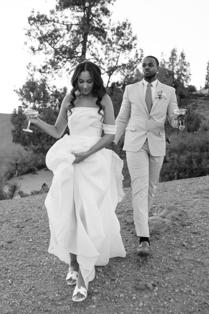 A bride and groom walk outdoors on a dirt path, holding drinks. The bride wears a flowing off-the-shoulder dress and sandals, while the groom wears a light-colored suit and tie. Trees and bushes are in the background.