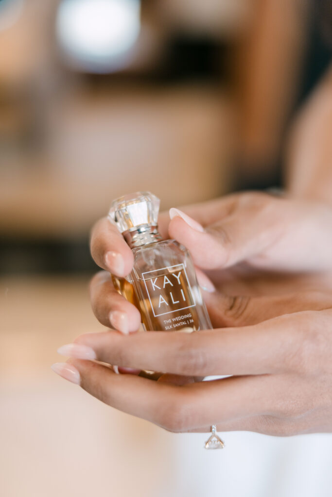 A person with manicured nails holds a small, elegant perfume bottle labeled KAYALI The Wedding Silk Santal 36. A delicate ring with a large stone is visible on one finger.