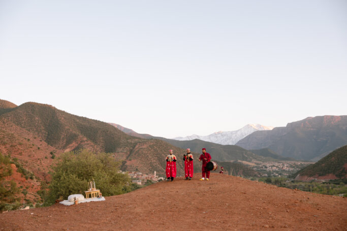 Three people in red traditional clothing stand on a hilltop overlooking a valley with mountains in the background under a clear sky. A small arrangement of objects sits on the ground to the left.