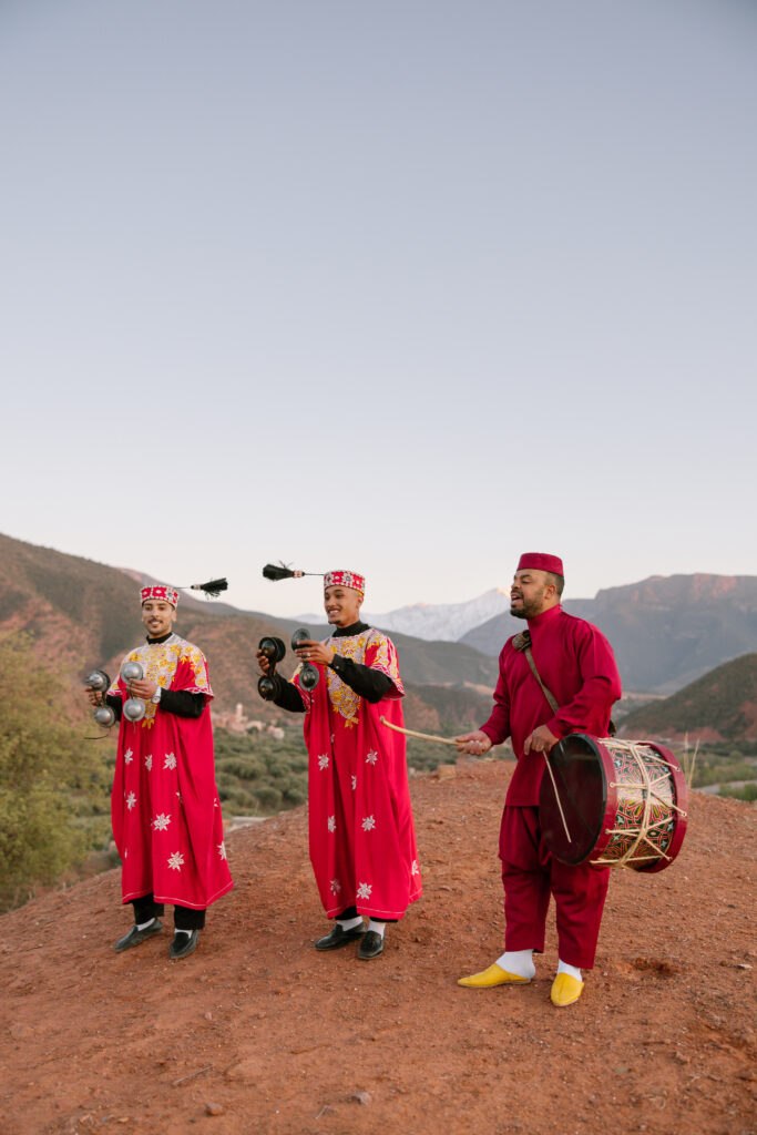 Three men in traditional red Moroccan attire perform music on a hillside; two play metal castanets while one plays a large drum. Snow-capped mountains and a clear sky are visible in the background.