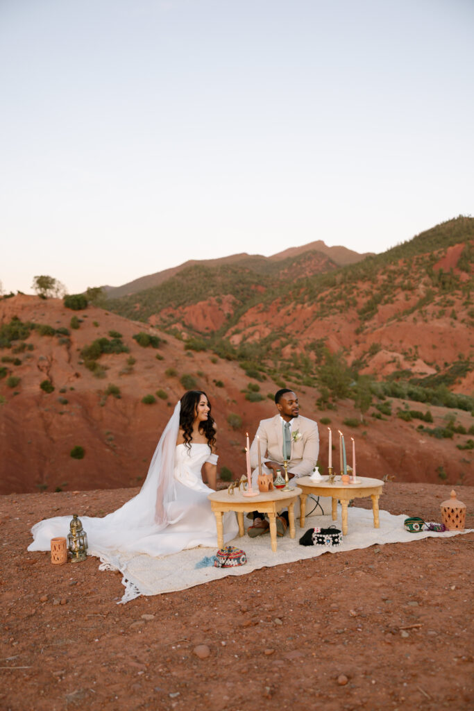 A bride and groom sit at a low table set for a romantic meal on a blanket outdoors, surrounded by candles and decor, with red rocky hills and mountains in the background.