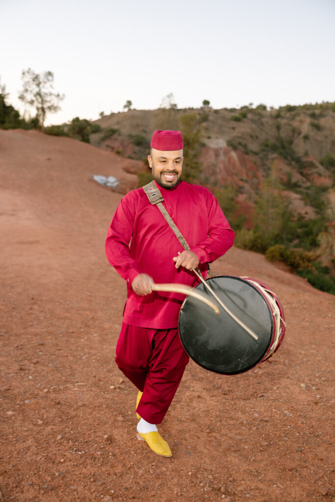 A man in a red traditional outfit and yellow shoes plays a large drum with sticks outdoors on a dirt path, with trees and hills in the background.