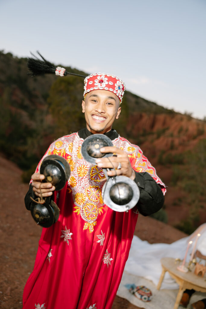 A smiling person in a bright red traditional robe and embroidered hat holds metal castanet-like instruments outdoors, with hills and a table with candles in the background.