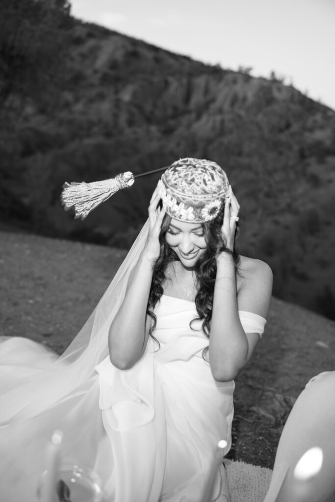 A woman in a strapless dress sits outdoors, smiling and adjusting a tasseled hat on her head. The background shows a blurred hillside. The image is in black and white.